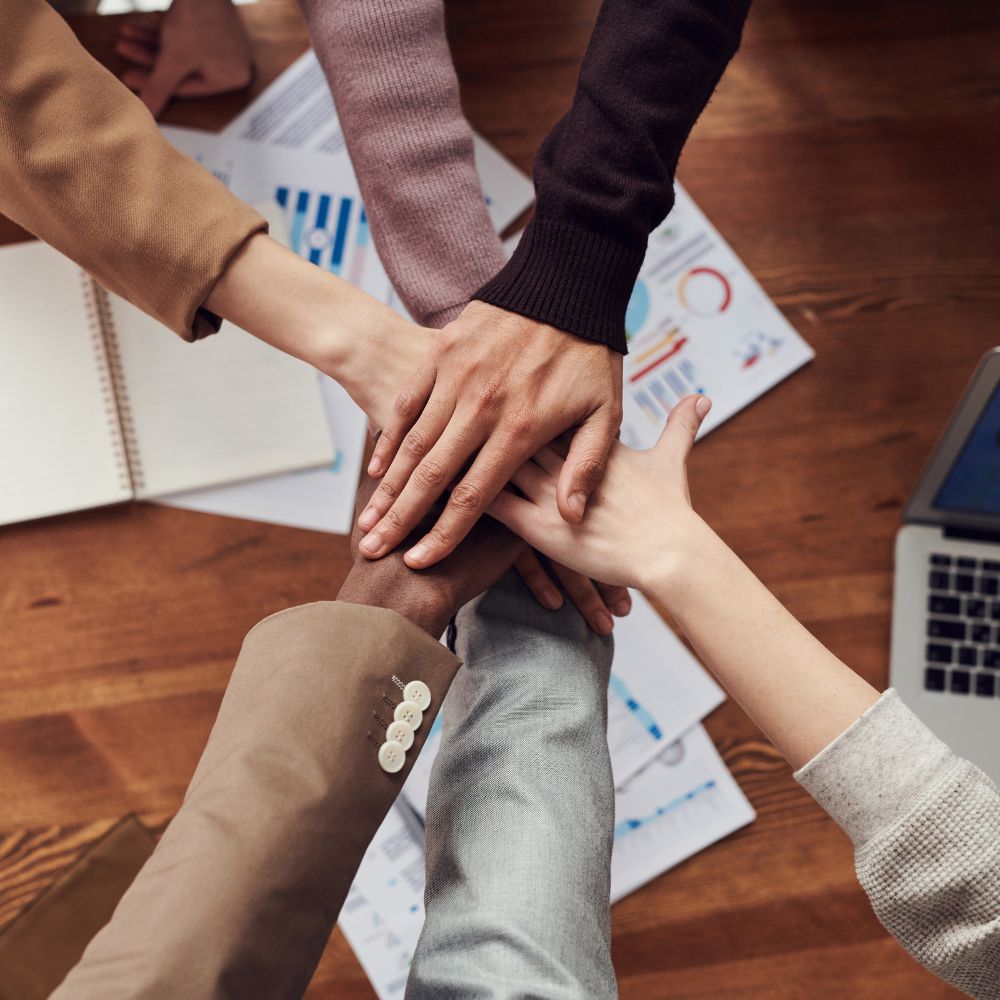 Four people stacking their hands together on a wooden desk with business materials.